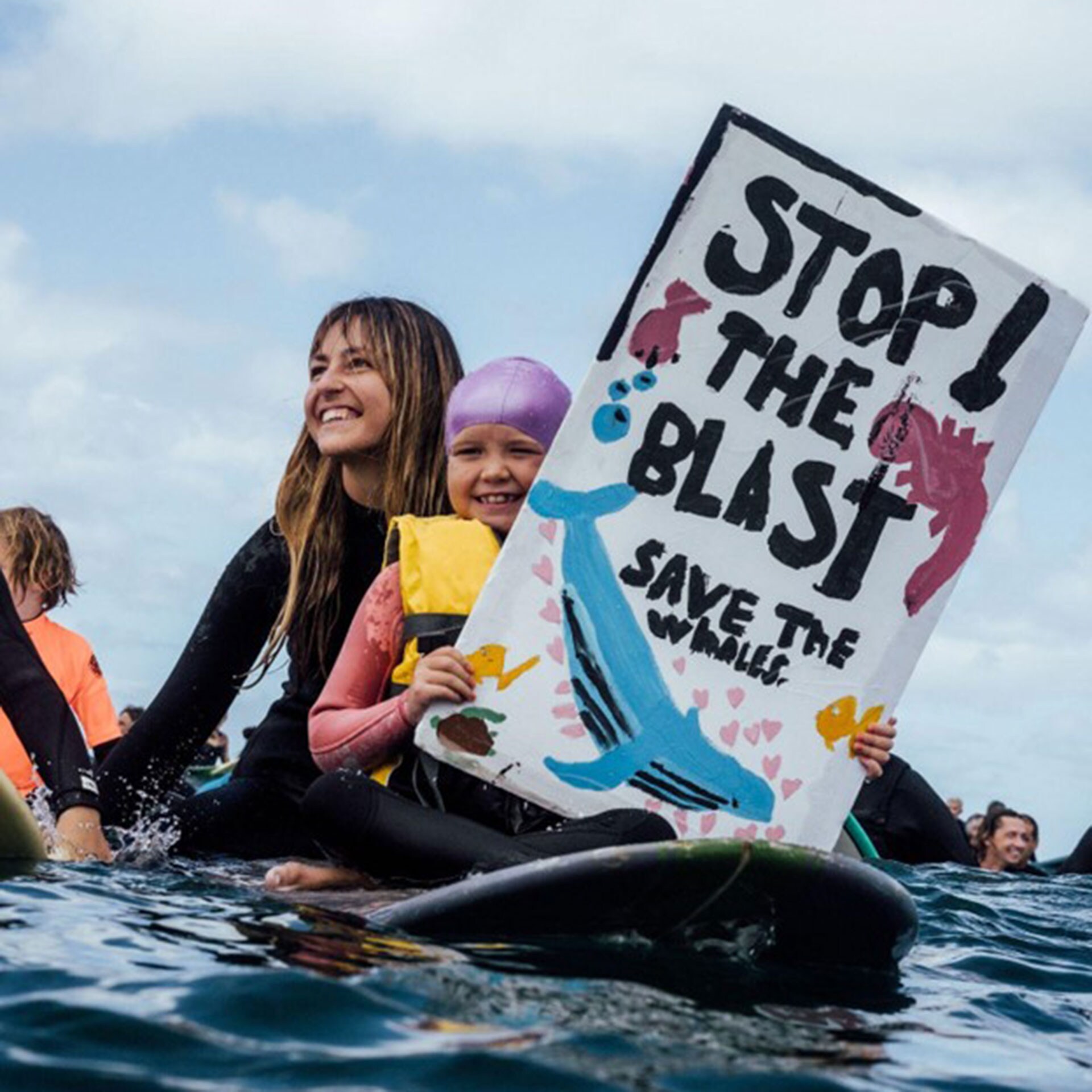 Two people floating on a surfboard holding a sign that reads 