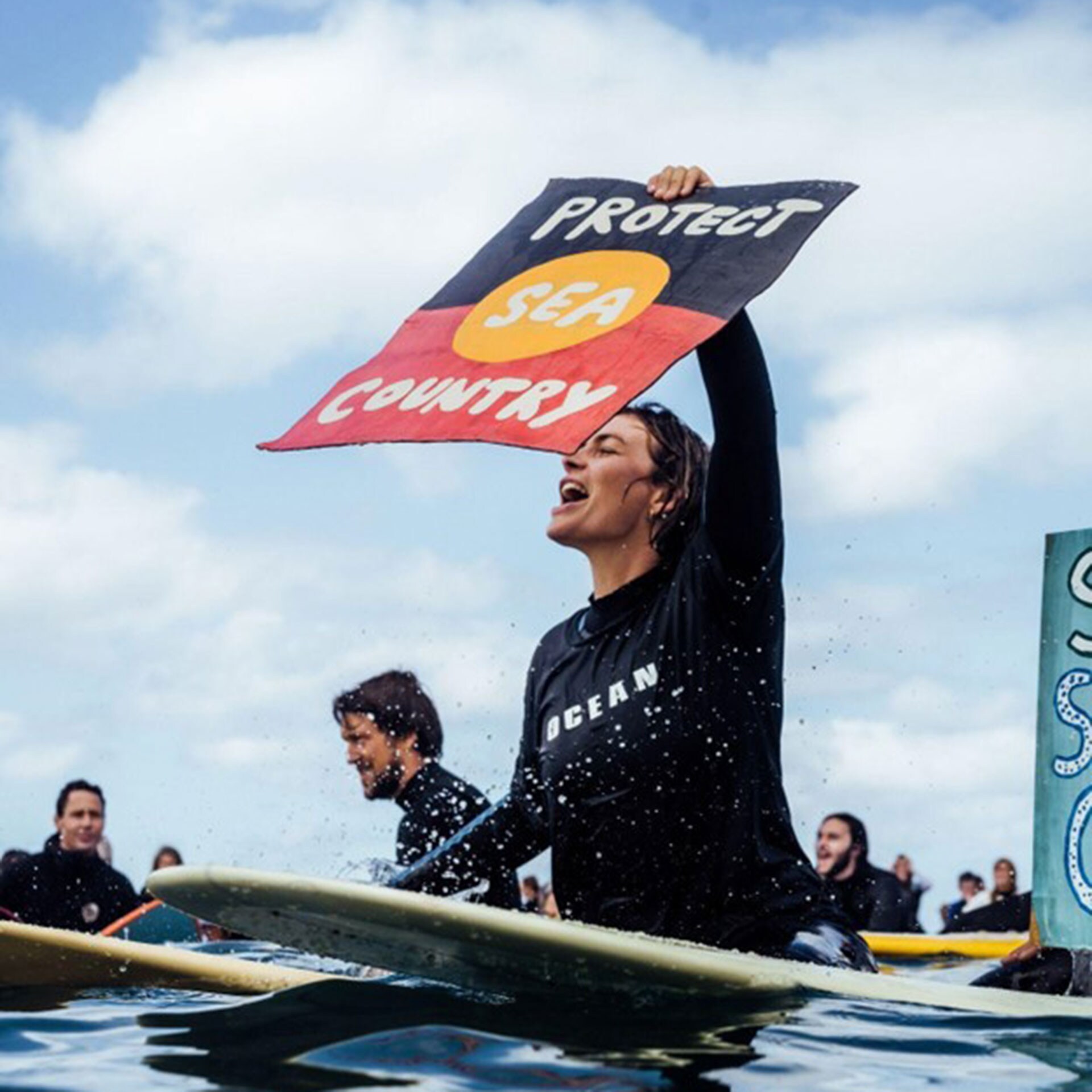 A person floating on a surfboard yelling and holding a sign that reads 