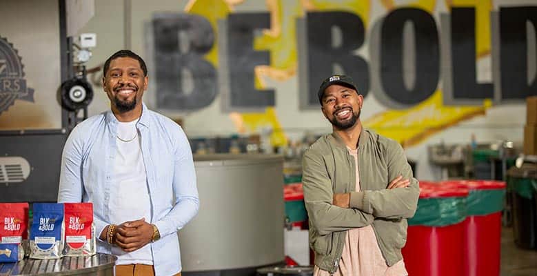 BLK & Bold co-founders Rod Johnson and Pernell Cezar stand in a coffee production facility with bags of BLK & Bold coffee.